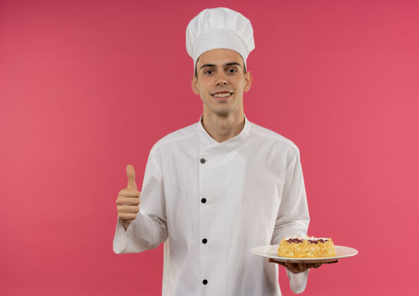 厨师面带微笑的年轻男厨师穿着厨师制服 大拇指举着盘子上的蛋糕戴着蛋糕拇指
