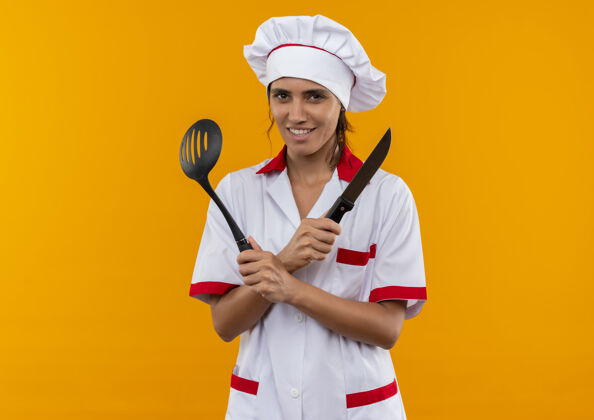 手持面带微笑的年轻女厨师穿着厨师制服拿着抹刀和刀子与副本空间女抹刀交叉