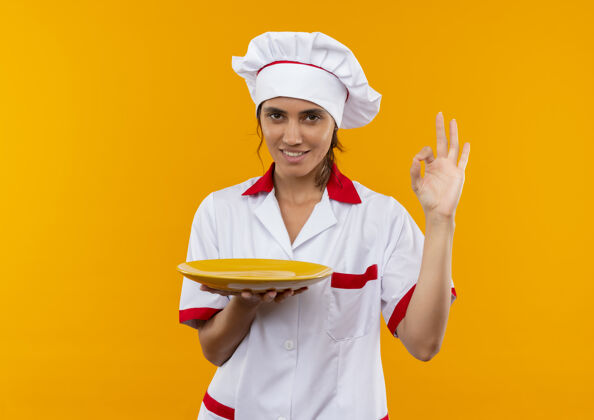 厨师面带微笑的年轻女厨师穿着厨师制服拿着盘子 展示着模仿空间的奥基手势好的穿着盘子