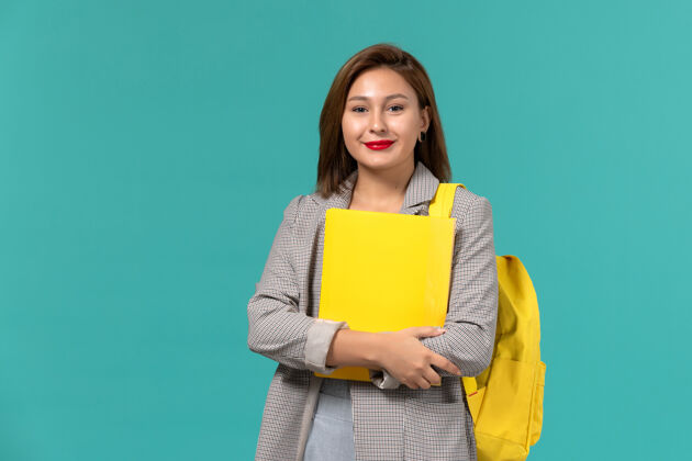 书身穿灰色夹克 背着黄色背包 在浅蓝色墙上拿着文件的女学生的正视图快乐背包灯