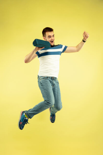 智能手机天空之声黄色背景的快乐跳跃人的全长肖像现代科技 自由选择的概念 情感的概念使用便携式扬声器像飞行中的超级英雄电话手机跳跃