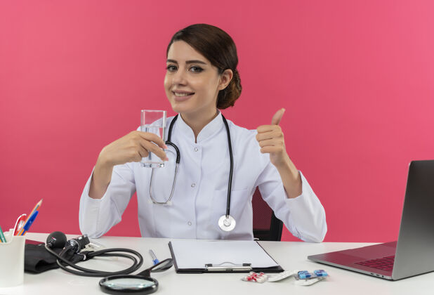 医疗面带微笑的年轻女医生穿着医用长袍 手持听诊器坐在办公桌旁 拿着医疗工具 拿着一杯水 拇指朝上 留着复印空间工具长袍拿着