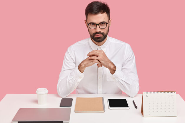 笔记本电脑自信帅气的留着胡须的年轻人穿着白色正式衬衫 戴着透明眼镜 坐在桌面上 完美主义者认真完美主义者记事本