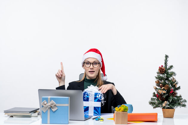 帽子圣诞节的心情与自信的年轻女子戴着圣诞老人的帽子和眼镜坐在一张桌子上拿着礼物 并显示在白色背景以上圣诞老人眼镜抱着