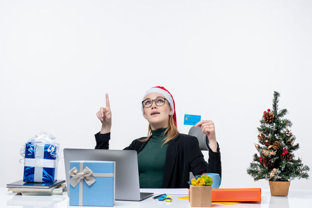 拿着一个戴着圣诞老人帽子 戴着眼镜 坐在桌边拿着圣诞礼物 手里拿着银行卡的漂亮女人指着办公室的上方笔记本电脑眼镜女人