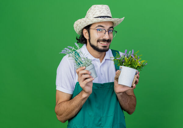 高兴年轻的留着胡子的园丁 穿着连体衣 戴着帽子 手里拿着盆栽植物 高兴地笑着年轻盆栽连身衣