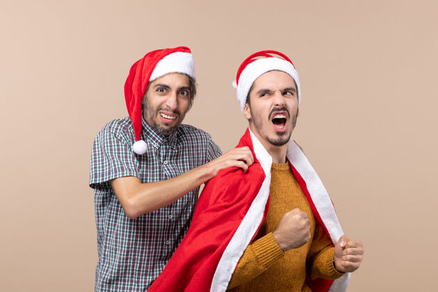 服装前视图两个圣诞节的家伙一个把圣诞老人外套放在他朋友的肩膀上米色的孤立背景朋友前面帽子