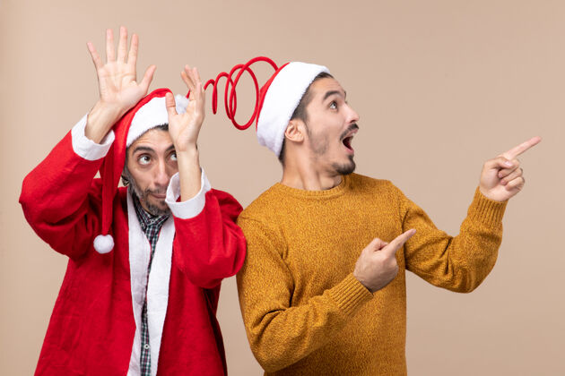 视图正面图：两个戴着圣诞帽的男人 一个把手举到头上 另一个在米色孤立的背景上显示方向男人肖像微笑
