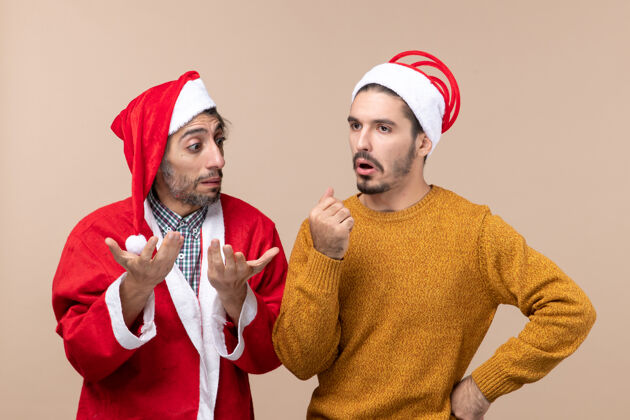 微笑正面图：两个戴着圣诞帽的男人在米色孤立的背景下思考着什么前面成人帽子