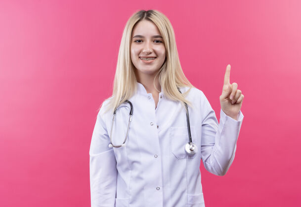 向上微笑的医生年轻的金发女孩穿着医用长袍戴着听诊器和牙套指着孤立的粉红色背景微笑穿着牙科
