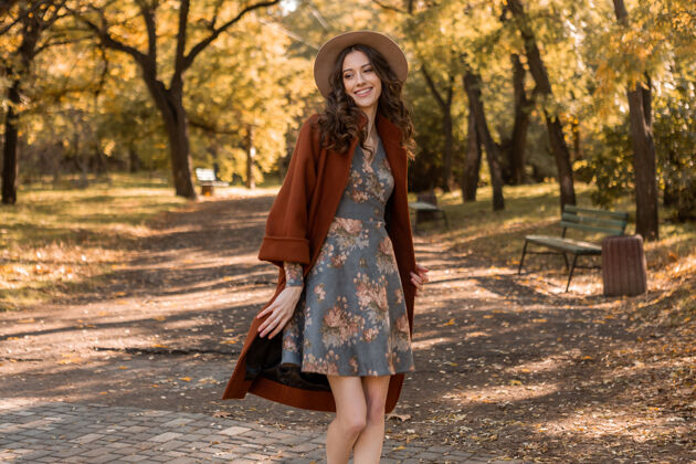 穿着迷人时尚微笑的女士 卷发漫步公园 身着印花连衣裙和保暖外套 秋季时尚 街头风格连衣裙波西米亚服装