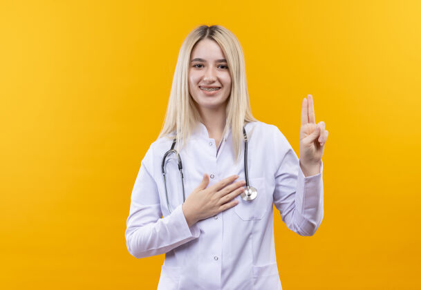 微笑微笑的医生年轻女孩穿着医用长袍戴着听诊器和牙套 在孤立的黄色背景上显示两个穿着两个医生