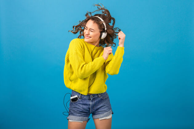 五颜六色迷人的笑容可掬的快乐女人戴着耳机跳舞听音乐 穿着时髦的服装 隔离在蓝色的工作室背景下 穿着短裤和黄色毛衣情感快乐兴奋