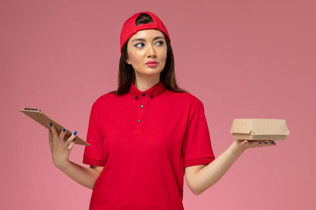 年轻女快递正面图：身穿红色制服斗篷的年轻女快递员 手上拿着小快递食品包和记事本放在粉红色的墙上服务工作漂亮