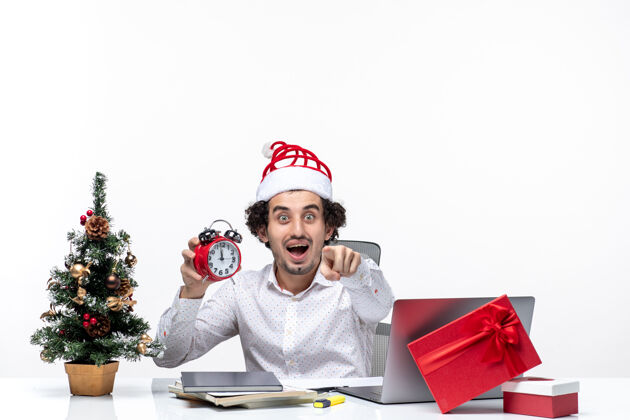 时钟年轻兴奋的商人戴着圣诞老人的帽子 在白色的背景下独自坐在办公室里看时钟圣诞老人帽子兴奋