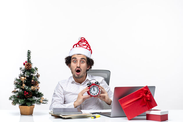人戴着圣诞老人帽 拿着闹钟 满脸胡须的年轻商人惊奇地看着什么东西 坐在白色背景的办公室里什么年轻圣诞节
