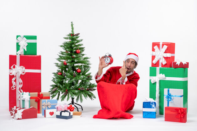 礼物情绪激动的圣诞老人惊讶地坐在地上 在礼物和装饰圣诞树旁的白色背景上展示钟表地面坐着礼物