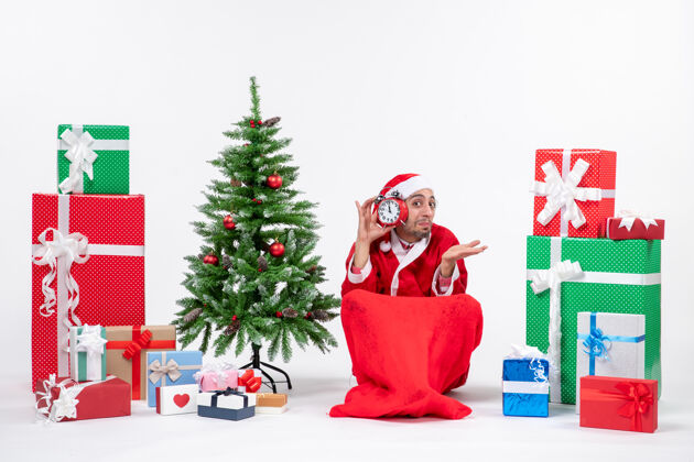 地情绪激动的圣诞老人惊喜地坐在地上 在礼物和装饰圣诞树旁的白色背景上展示钟表礼物情绪化惊讶