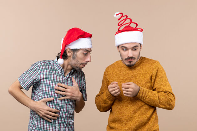 视图前视图两个戴着圣诞帽的困惑的家伙一个看着另一个手放在米色孤立的背景上圣诞老人两个男人