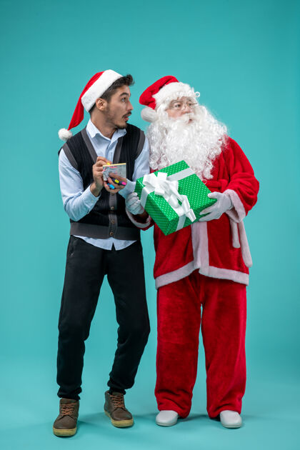 采取前视图：圣诞老人和正在蓝色背景上做笔记的年轻男性视图圣诞老人圣诞