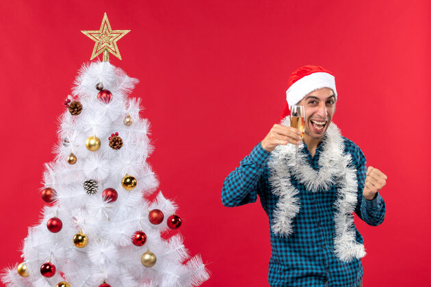 圣诞树圣诞节心情与情绪激动的年轻人圣诞老人帽子在一个蓝色条纹衬衫举起一杯葡萄酒附近圣诞树搞笑年轻人小伙子