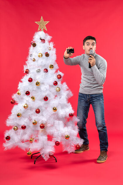 站立圣诞气氛 骄傲的家伙站在装饰好的圣诞树旁 拿着麦克风和手机展示着什么东西心情装饰