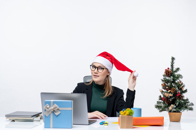 笔记本电脑新年气氛积极的金发女人 戴着圣诞老人的帽子 坐在一张桌子旁 桌子上放着一棵圣诞树和一份白色背景的礼物圣诞节圣诞老人女商人