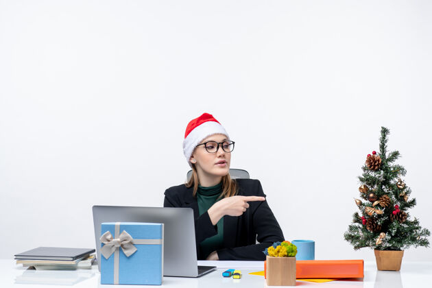 漂亮的东西新年气氛与年轻迷人的女人坐在一张桌子上圣诞老人帽子圣诞树和礼物 它指向办公室的东西指着电脑桌子