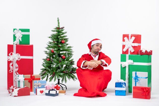 圣诞老人惊讶激动的年轻人打扮成圣诞老人 拿着礼物和装饰好的圣诞树坐在白色背景的地上打扮坐着圣诞树