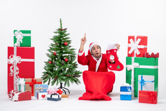 袜子严肃的圣诞老人指着上面坐在地上 拿着圣诞袜靠近礼物和装饰在白色背景上的新年树拿着圣诞老人圣诞老人