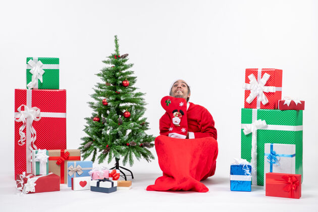 圣诞搞笑的圣诞老人坐在地上 穿着圣诞袜靠近礼物和装饰圣诞树的白色背景股票照片圣诞老人礼物圣诞老人