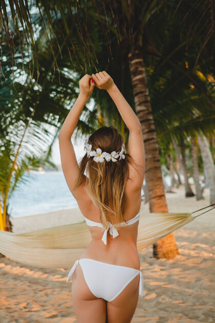 海洋穿着白色比基尼泳装的年轻性感女人在热带海滩 棕榈树 夏威夷摆姿势 头发上的花朵 性感 苗条的身体 阳光明媚 从后面看 享受假期 在岛上旅游人身体乐趣