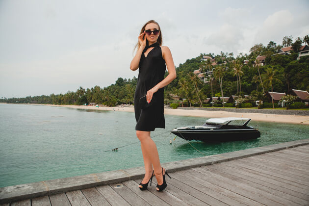 海洋奢华性感迷人的女人穿着黑色礼服在豪华度假酒店的码头上摆造型 戴着墨镜 暑假 热带海滩异国情调船游艇