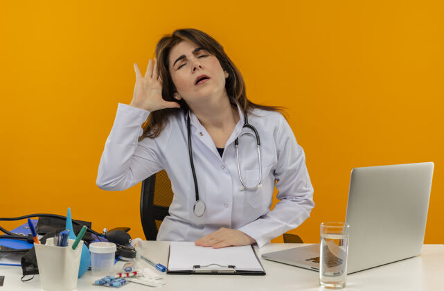 长袍疲惫的中年女医生穿着医用长袍 带听诊器 坐在办公桌旁 用笔记本电脑和医疗工具 手放在头上 孤立的橙色背景上 有复印空间工具放女