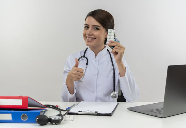 工作面带微笑的年轻女医生穿着医用长袍 手持听诊器坐在办公桌旁 拿着医疗工具 拿着药片 拇指朝上 留着复印空间电脑医生药片