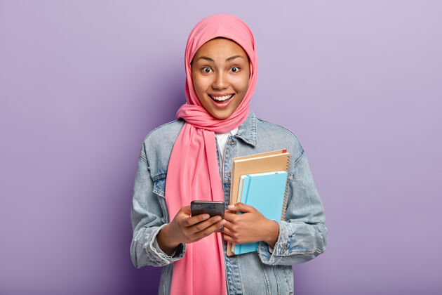 高兴快乐的妇女手握笔记本和手机 头上戴着粉色面纱手机设备阿拉伯语