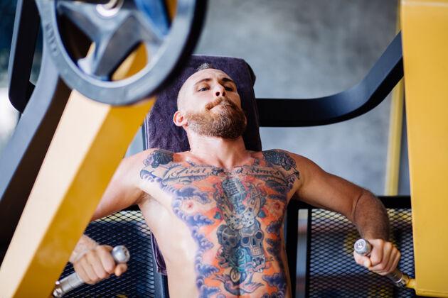 二头肌在靠近窗户的健身房里 纹身的大胡子男人在胸部按压机上锻炼的肖像体重人健美