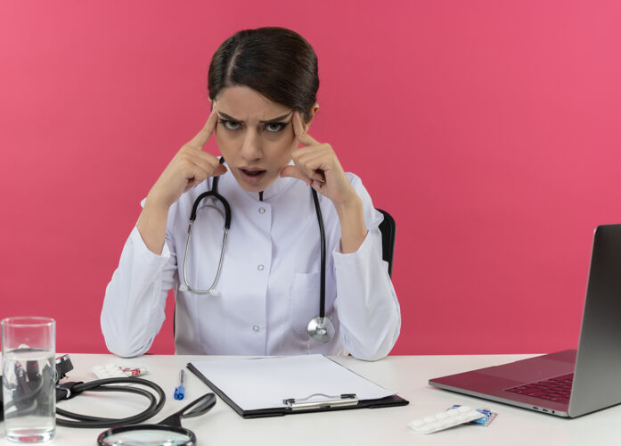 放困惑的年轻女医生穿着医用长袍 手持听诊器坐在办公桌旁 拿着医疗工具 把手指放在头上 放在隔离的粉色墙壁上 留着复印空间坐着医生头部