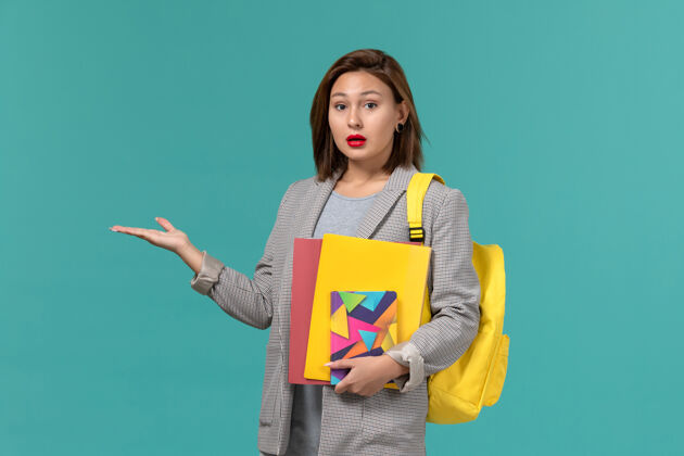 Female身穿灰色夹克 背着黄色背包 在蓝色墙上拿着文件和文案的女学生的正面图背包字帖夹克