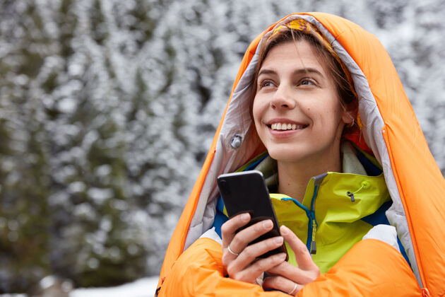 短信快乐的女游客在睡袋里取暖 在积雪覆盖的山顶上摆姿势 手持现代手机科技山脉冒险