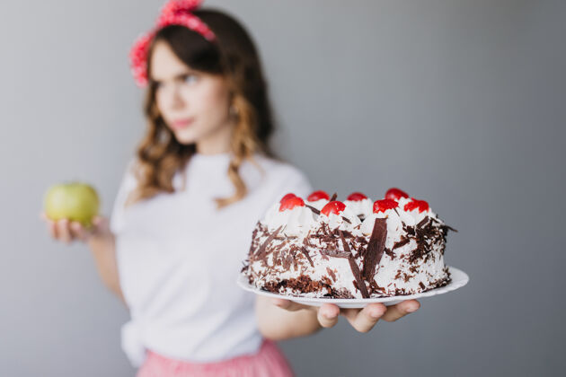 休闲端详优雅的女孩 手里拿着巧克力蛋糕 想着饮食模糊的黑发女人的肖像 前景是奶油馅饼蛋糕苹果巧克力