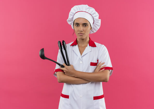 厨师年轻的女厨师穿着厨师制服 拿着勺子 双手交叉放在隔离的粉色墙壁上 还有复制空间手看年轻