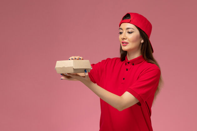人正面图：身穿红色制服斗篷的年轻女快递员 手上拿着一个小小的快递食品包 放在淡粉色的墙上送货快乐服务