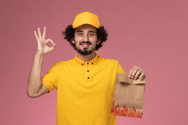 工作正面图穿着黄色制服的男快递员拿着浅粉色墙上的纸质食品包男工人正面