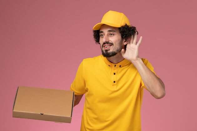 工人正面图身穿黄色制服的男快递员拿着食品快递箱 试图听到淡粉色墙上传来的声音披萨人人
