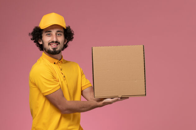 快餐正面图：身穿黄色制服的男快递员拿着食品快递箱站在浅粉色的墙上持有工作快餐
