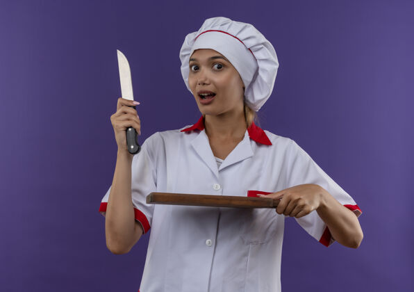 惊讶惊讶的年轻厨师女身穿厨师制服手持刀和砧板就孤立的背景拿着板子穿着