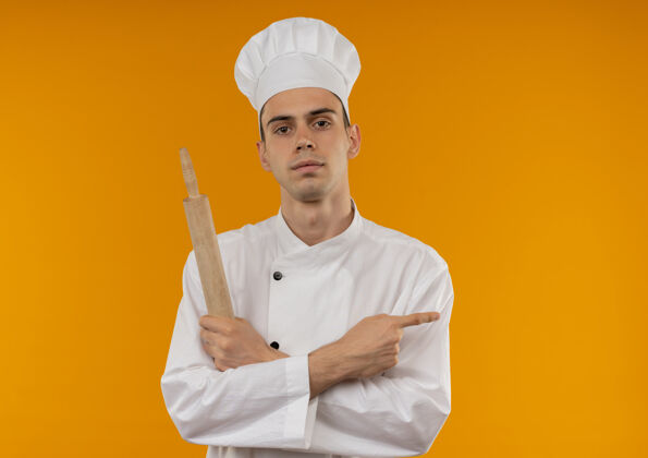厨师年轻男酷穿厨师制服交叉手拿擀面杖手指指向一边孤立的黄色墙壁与复制空间穿交叉侧
