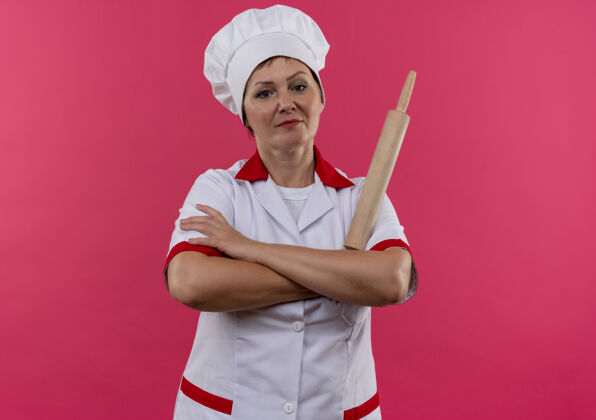 拿着自信的中年女厨师穿着厨师制服 手拿擀面杖交叉在孤立的粉红色墙上手滚动厨师