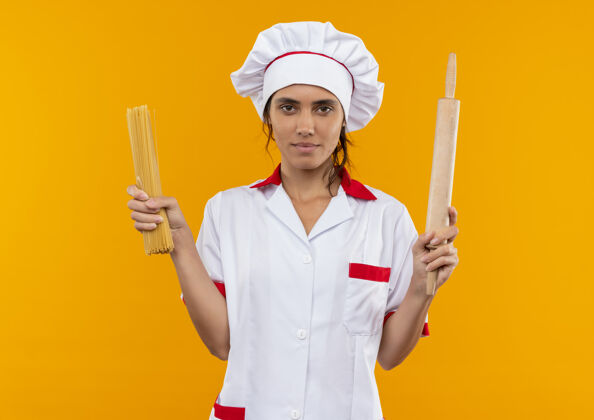 拿着看起来年轻的女厨师穿着厨师制服拿着意大利面和擀面杖站在隔离的黄色墙壁上 有复制空间意大利面滚看